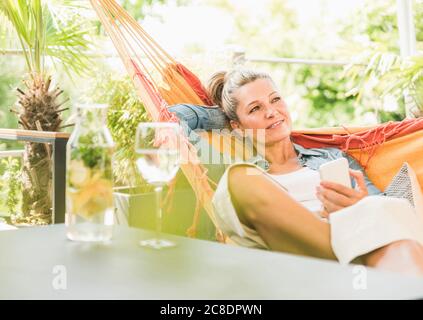 Portrait of pensive mature woman with smartphone relaxing in hammock on terrace Stock Photo