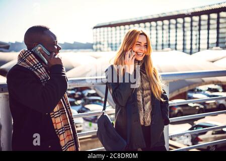 Cheerful colleagues talking over smart phone while standing by railing on bridge at station Stock Photo