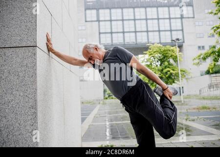 Senior man stretching leg while standing by wall on footpath in city Stock Photo
