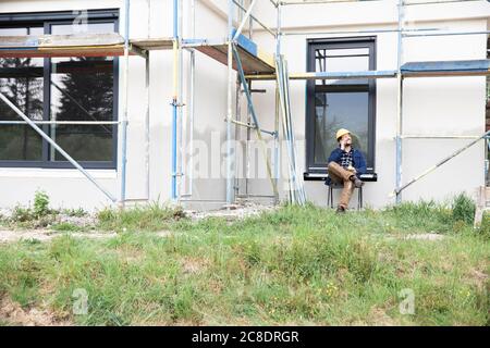 Construction worker talking over mobile phone while sitting against house Stock Photo