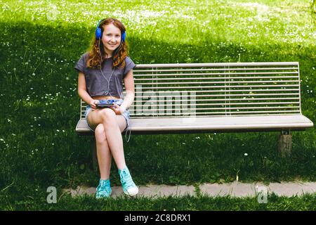Smiling girl listening music through headphones while sitting on bench at park Stock Photo