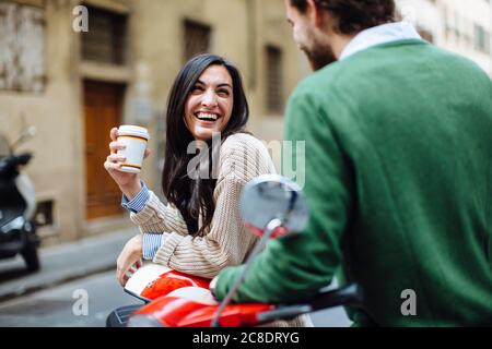 Happy woman holding coffee looking at boyfriend while leaning on Vespa Stock Photo