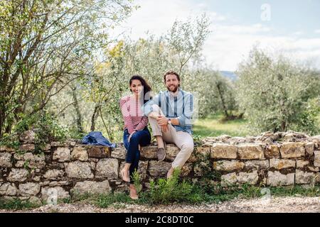 Smiling young couple sitting on retaining wall against trees Stock Photo
