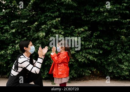 Mother and daughter wearing masks while playing clapping game against plants Stock Photo