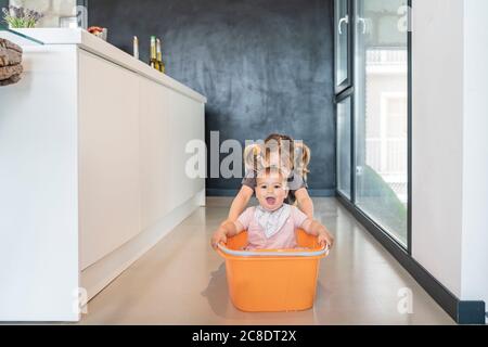 Girl pushing baby sister sitting in bucket on floor in modern kitchen Stock Photo