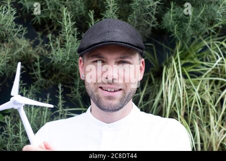 Smiling male entrepreneur wearing cap holding wind turbine model against plants in park Stock Photo