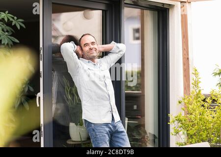 Smiling mature man leaning on window with hands behind head at back yard Stock Photo