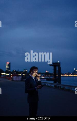 Mature businessman using tablet and smartphone at the riverbank at dusk Stock Photo
