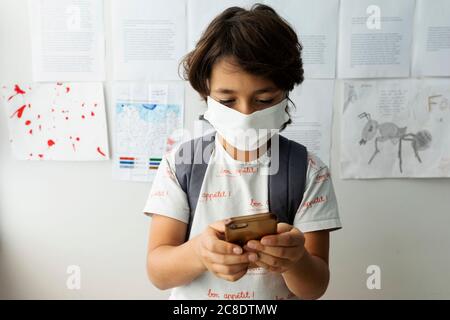 Boy wearing mask using smart phone while standing against wall in school Stock Photo