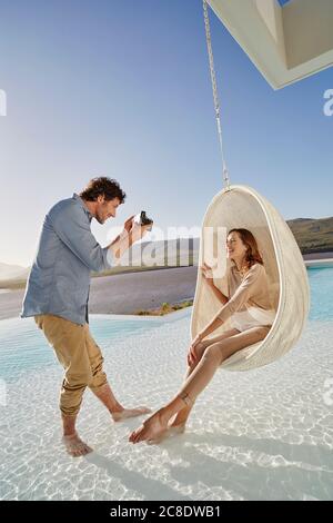 Man taking picture of woman sitting in hanging chair above swimming pool Stock Photo