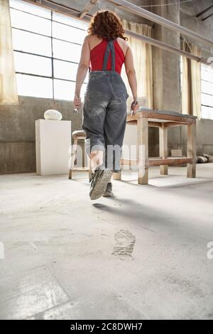 Woman wearing overalls walking on dusty floor towards table in workshop Stock Photo