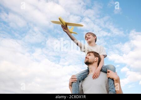 Man carrying son on shoulders playing with toy airplane against sky Stock Photo