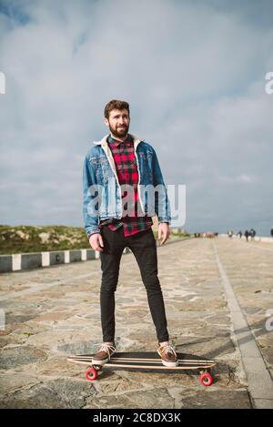 Handsome bearded man skateboarding on footpath against cloudy sky in park Stock Photo