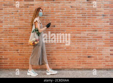 Young woman wearing mask using smart phone while walking on footpath by brick wall Stock Photo