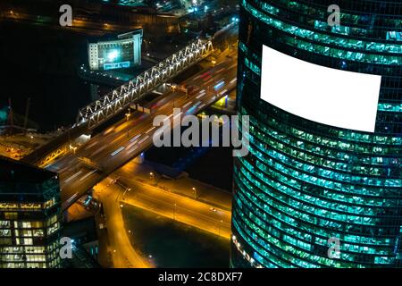 Mock up image: blank white billboard or large display on skyscraper, fast moving cars traffic and warm street light at night. Mockup, advertising Stock Photo