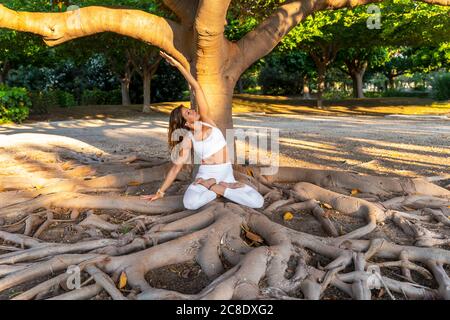 Mature woman with arm raised exercising while sitting on roots in park Stock Photo