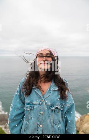 Smiling female cancer survivor woman with eyes closed standing against sea and cloudy sky Stock Photo