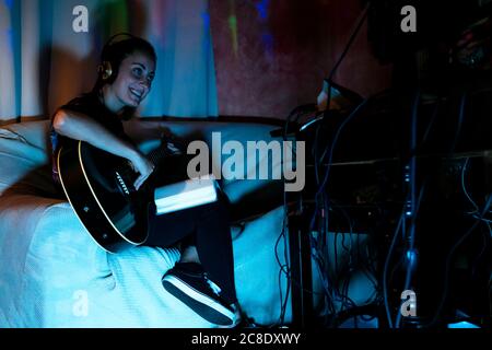 Happy female musician playing guitar on sofa in recording studio at home Stock Photo