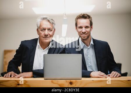 Two confident businessmen sitting in office, using laptop, smiling Stock Photo