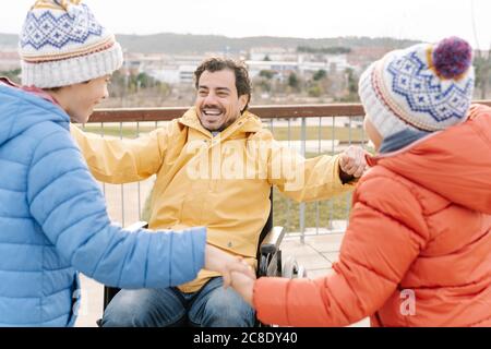 Cheerful father sitting on wheelchair holding sons hands while playing in park Stock Photo