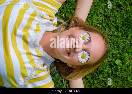 Close-up of smiling woman with oxeye daisies on eyes lying over grassy land in park Stock Photo