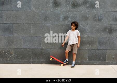 Boy playing with skateboard while standing on footpath against wall Stock Photo