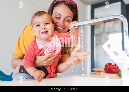 Smiling mother and cute baby girl playing with water falling from faucet in kitchen sink Stock Photo