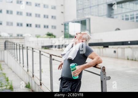 Tired senior man holding bottle yawning while standing by railing in city
