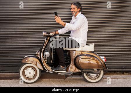 Happy mature man taking selfie while sitting on motor scooter against black shutter Stock Photo