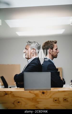 Two confident businessmen sitting in office with laptop, back to back Stock Photo