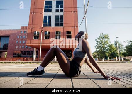 Thoughtful female athlete listening music through headphones while sitting on street in city Stock Photo