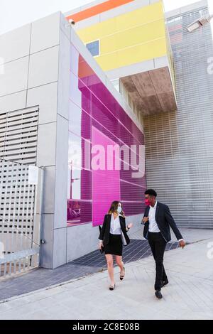 Colleagues wearing masks talking while walking on footpath against building in city Stock Photo