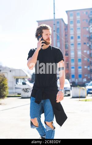 Handsome man talking over smart phone while walking on street during sunny day Stock Photo