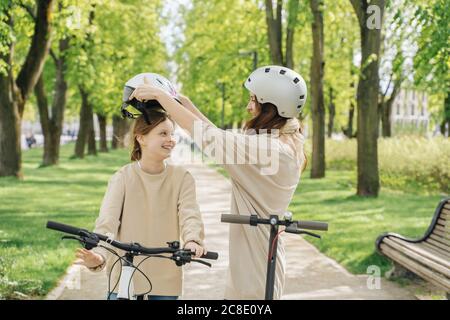 Mother wearing helmet to her daughter while standing against trees in park Stock Photo