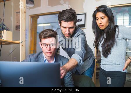 Three business people sharing laptop at desk in office Stock Photo