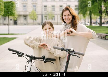 Mother with daughter gesturing heart shapes while standing by bicycle and scooter in city park Stock Photo