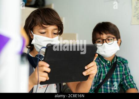 Boy wearing face mask using digital tablet while sitting in distance with friend at school Stock Photo