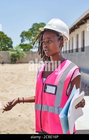 Female architect holding papers looking away while standing at construction site during sunny day Stock Photo