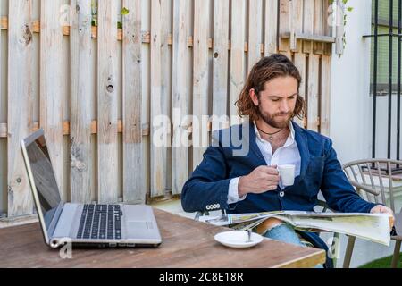 Handsome young businessman with long brown hair reading newspaper while drinking espresso coffee at cafe Stock Photo