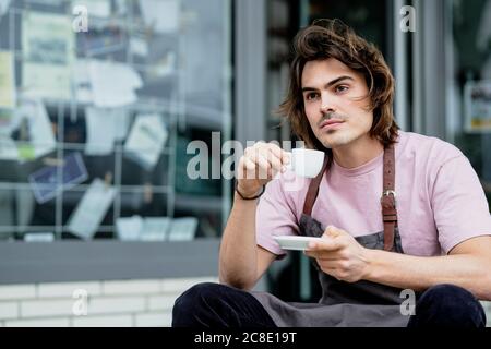 Thoughtful male barista holding coffee sitting outside coffee shop Stock Photo