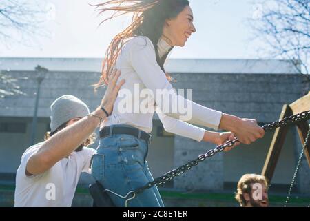 Young man pushing playful friends swinging in park during sunny day Stock Photo
