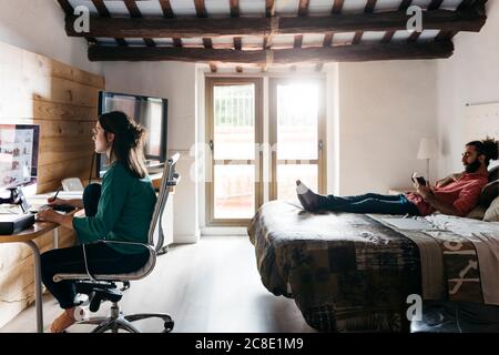 Female architect using computer at home, man reading a book lying on bed Stock Photo