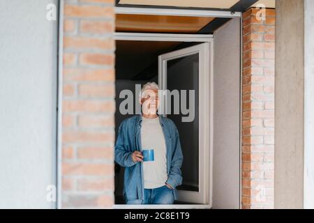 Thoughtful senior man holding coffee mug while looking through window Stock Photo
