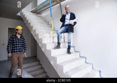 Male architect talking over smart phone while construction worker working in house under construction Stock Photo