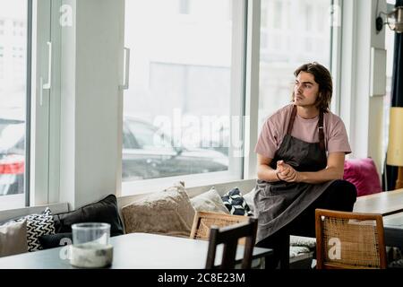 Thoughtful barista with hands clasped sitting on table in cafe Stock Photo