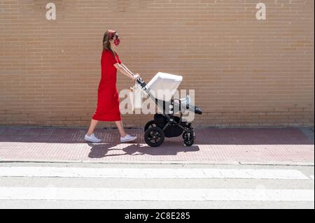 Mother wearing mask pushing son in baby carriage while walking on sidewalk Stock Photo