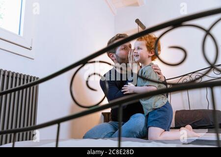 Father whispering to son on bed at home Stock Photo
