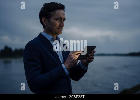 Mature businessman using tablet at the riverbank at dusk Stock Photo