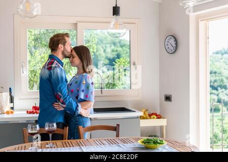 Man kissing woman while standing by dining table in kitchen at home Stock Photo