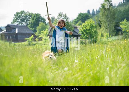 Playful boy wearing cape running with dog on grassy land Stock Photo
